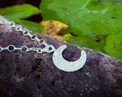 Silver necklace with a crescent moon pendant on a stone surface with green leaves in the background