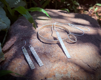 Silver jewelry set including a necklace, bracelet, and earrings on a stone surface with green foliage in the background.